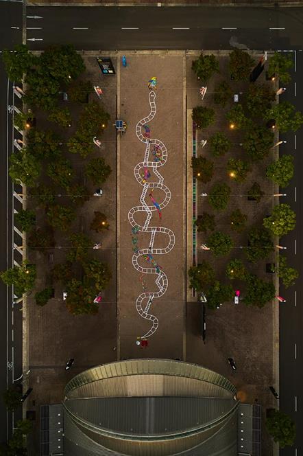 An aerial view of a mural painted on the ground. The mural is a giant snakes and ladders board.