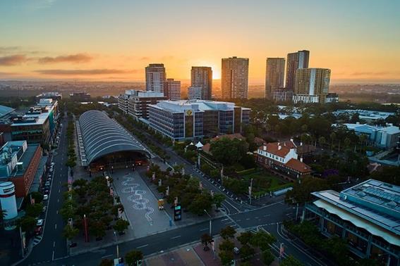 Aerial view of the artwork Snakes and Ladders, and the surrounding suburb of Sydney Olympic Park. The artwork is in a large paved space outside a train station building with an arched roof.