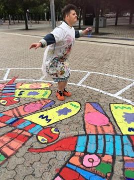 Digby is wearing a white smock covered in colourful paint, crouching amidst colourful dragonflies painted on the ground. He is pretending to fly like a dragonfly.