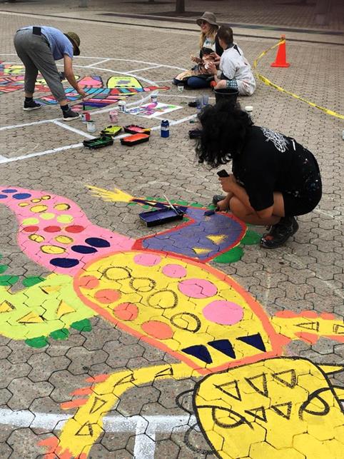 Four people are painting the mural on the ground. They are working with paint trays and rollers, and many colours.