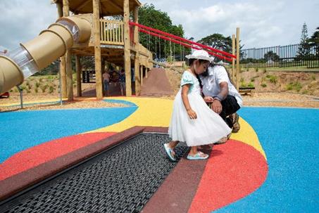 A girl with caregiver stepping off the ground level trampoline that is integrated with the vibrant coloured rubberised matting. In the background is the inclusive slide.
