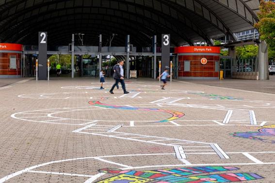 Two adults and two children are playing on the Snakes and Ladders artwork. A colourful snake stretches across the middle of the artwork.