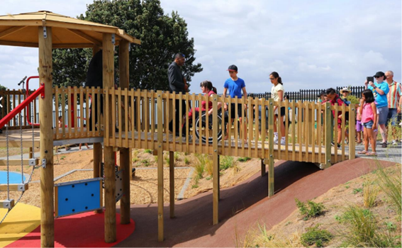 Keith Park playground's inclusive slide with level entry ramp being used by a child in wheelchair with caregivers and other children also using the ramp. 