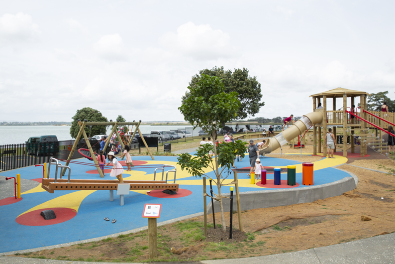 Wide angle view of the redeveloped Te Pua/Keith Park. In the foreground is the inclusive see-saw and in ground trampoline. In the background are the swings including the basket swing and caregiver and child swing. To the right is the wheelchair accessible play tower reached by a gentle gradient ramp. The playground surface is colourful blue red and yellow, guiding people to the different play features.