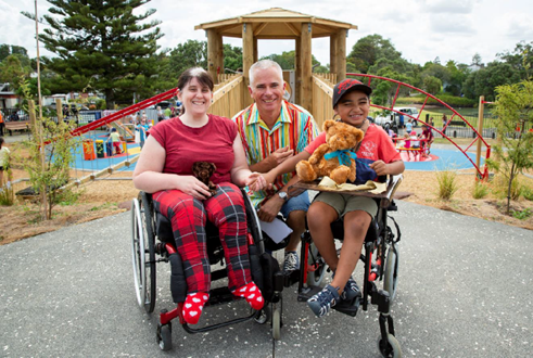 Pictured are two new playground users Emma and Israel who both use wheelchairs for their mobility. They are holding hands and have teddy bears on their laps. Pictured crouching behind them is local board chair Joseph Allan. All are smiling. In the background is the new customized 2m high wheelchair accessible play tower.